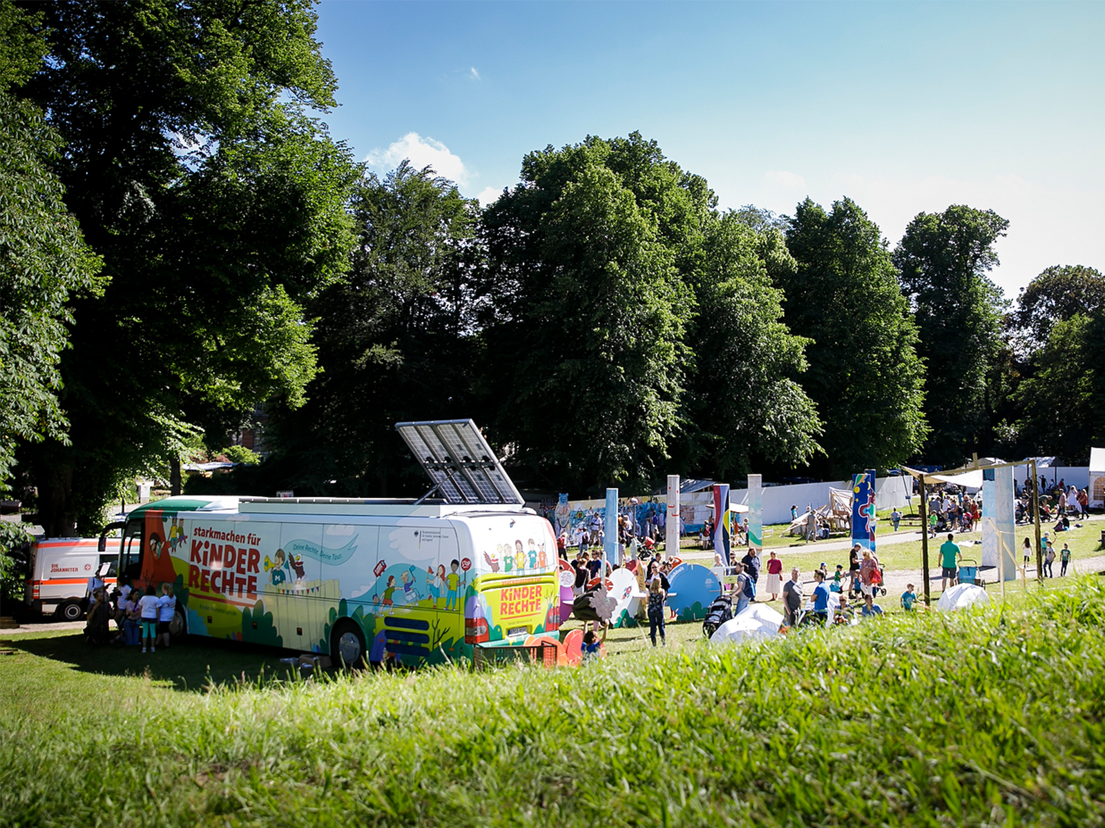 Der Kinderrechte-Bus steht auf einer großen, hügeligen Wiese, auf denen die verschiedenen Stationen zu den Kinderrechten aufgebaut sind.